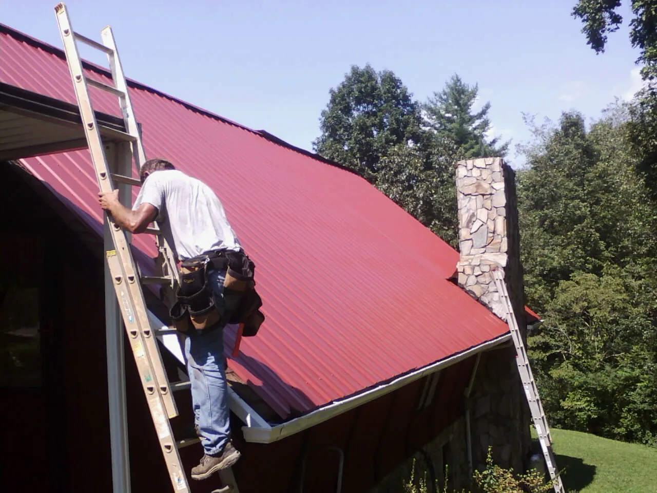 Roofer on a ladder beside a red metal roof and stone chimney — Soffit & Fascia Repair by Roofing Brothers in East Falmouth