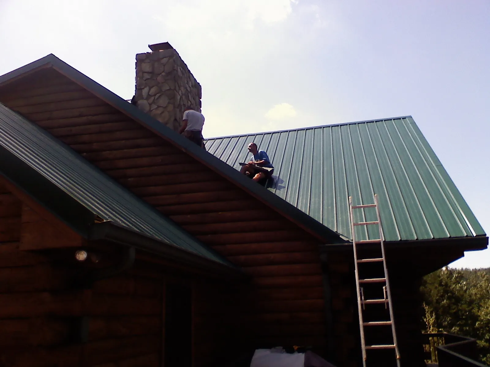 Roofers installing green metal panels beside a stone chimney on a log home in East Falmouth